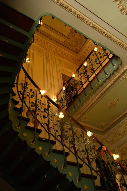 A view looking up a decorated Victorian staircase during a home relocation in NW3, with ornate gold and cream ceiling details and intricate wrought iron balustrades. The staircase has dark wooden handrails, and the steps are green with decorative star-shaped embellishments on the risers. Warm lighting from small chandeliers illuminates the staircase, as two people are seen carrying a large, bulky wooden wardrobe wrapped in protective padding along the staircase for safe furniture transport. The setting suggests a professional removals process by Man with Van Primrose Hill, ensuring safe packing and moving of significant household furniture through a historic property during an interior house move.