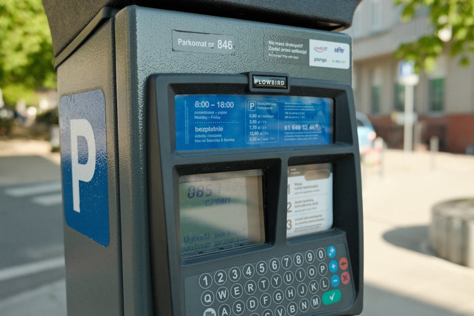 Close-up view of a parking meter situated on a city street, with a blue sign displaying a white 'P' symbol indicating parking availability. The parking meter features a digital screen showing parking fee information and a smaller display showing a number, with a keypad below for inputting payment details. Surrounding objects include a curb and pavement, with a blurred background of residential buildings and greenery, illustrating an outdoor setting typical for house removals or furniture transport in Primrose Hill. The lighting suggests daytime, and the scene reflects the typical process of parking management for home relocation or moving services provided by Man with Van Primrose Hill, highlighting the importance of parking permits for efficient furniture transport and packing during house removals.