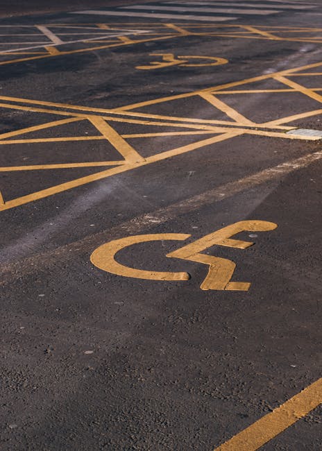 A close-up view of an outdoor parking area pavement with yellow painted markings, including a prominent wheelchair accessible parking symbol near the foreground. In the background, there are several other parking space lines and additional yellow markings, some of which are partially visible. The surface of the asphalt appears slightly worn with small patches and cracks, indicating regular use. The lighting is natural, likely daylight, providing clear visibility of the markings. This setting is typical for a residential or public parking lot where vehicle access for home relocation or furniture transport via professional removals services, such as those offered by Man with Van Primrose Hill, would take place during a home move or furniture delivery.