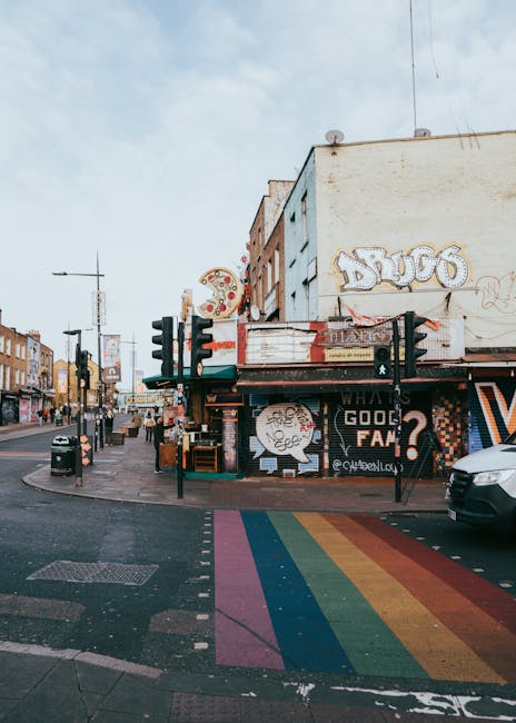 A busy urban street scene in Primrose Hill, NW1, showcasing a building with various graffiti and signage above a ground-floor shop. The shop appears to be a local eatery or bar, with an awning, outdoor display, and neon signs. Traffic lights and pedestrian crossing signals are positioned at the corner, with a black car parked on the right side near a rainbow-colored crosswalk painted on the pavement. The street includes several other buildings in the background, featuring brick and stucco facades, with signage for different businesses. Street furniture such as a black trash bin and bollards are visible along the sidewalk. The lighting suggests a daytime setting with partly cloudy skies. This scene exemplifies typical street activity in Camden, an area known for vibrant street art and community engagement, relevant to the context of local planning regulations like loading bay permits. Man with Van Primrose Hill, a removals service, may utilize such streets for home relocation and furniture transport, involving careful planning around urban features.