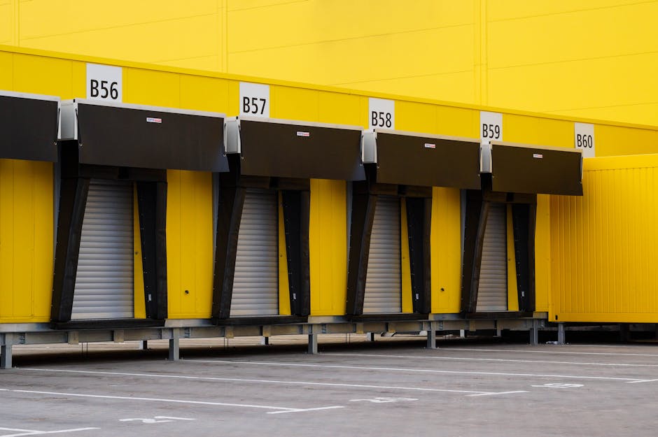 The image shows a large yellow commercial building with four truck bays, each labeled with white tags marked B56, B57, B58, B59, and B60. The bays are equipped with black retractable dock levelers folded down, used for loading and unloading goods during home relocation or furniture transport. The surface in front of the bays is a paved lot with white parking lines, indicating designated space for delivery vehicles. The bright yellow colour of the building contrasts with the grey pavement and the black dock equipment, highlighting the loading area designed for logistics and moving operations. This scene captures a typical industrial or warehouse environment where professional removals services, such as those offered by Man with Van Primrose Hill, commonly operate to facilitate home removals and packing and moving processes, potentially involving the use of loading bays and equipment in a controlled environment.