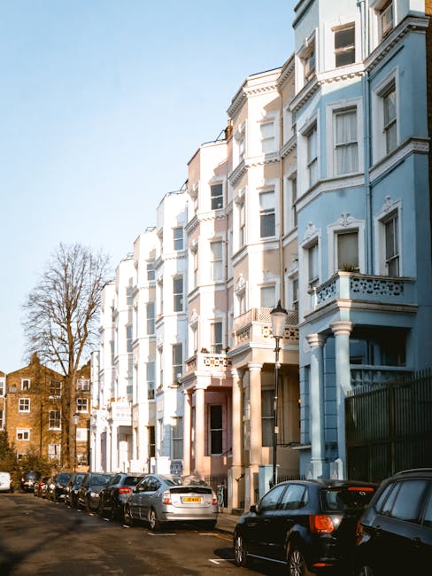 Exterior view of a multi-storey residential building with a light blue façade and decorative white architectural details, situated along a street lined with parked cars. The building features several bay windows with white frames, small balconies with ornate balustrades, and a main entrance with a brown door. Sunlight illuminates the building, casting shadows on the pavement, with a clear blue sky overhead. In the foreground, a row of vehicles, including sedans and hatchbacks, are parked along the curb on the paved street, which appears to be part of a residential area in Primrose Hill. The image also shows some leafless trees in the background, indicating a season of late autumn or winter. This scene is typical of a city neighbourhood where house removals and furniture transport by [COMPANY_NAME] may involve navigating street congestion and parking restrictions during efficient home relocation processes, especially in flats without lifts requiring careful planning for the loading process.