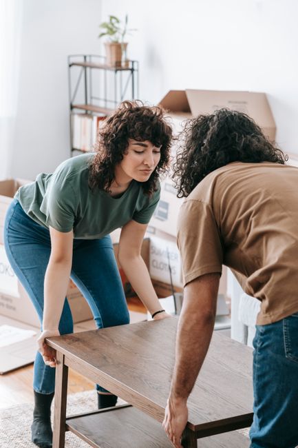 Two individuals are engaged in a home relocation task inside a well-lit room with a modern, minimalist decor. The woman, with short curly hair and wearing a green T-shirt and blue jeans, is carefully lifting a wooden table while the man, with longer curly hair and dressed in a brown T-shirt, is supporting the other side of the table. The table appears to be part of the furniture being prepared for transport. In the background, there is a tall metal shelving unit with multiple shelves holding potted plants, books, and storage boxes, along with a partially visible cardboard box and a large open cardboard box with additional packing materials nearby. The scene is set in an interior space with natural lighting, indicating the initial stages of a packing and furniture moving process, typical of house removals services offered by Man with Van Primrose Hill. The key elements include the cardboard packing boxes, wooden furniture, and careful handling during a furniture transport operation as part of a home relocation effort.