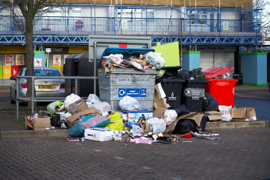 A collection of overflowing waste and recycling bins positioned on a paved sidewalk outside a commercial building with a construction scaffold in the background. The waste includes open cardboard boxes, plastic bags, and various packaging materials spilling onto the ground around the bins, which are mounted on a metal railing. The recycling containers are labeled for paper and mixed waste, while other bins are black and red for general waste. Several parked cars are visible behind the bins, and the environment is illuminated by daylight. This scene depicts the importance of proper waste disposal during home relocation and moving processes, as managed by professional removals services like Man with Van Primrose Hill.