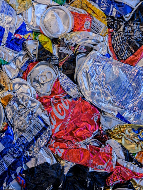 A close-up view of a pile of crushed and crumpled aluminium cans, including brands such as Coca-Cola and other soft drink containers, with various colours of metallic silver, red, blue, black, and gold packaging. Some cans are partially flattened with visible pull-tab tops, and others have remnants of labels. The cans are stacked irregularly, occupying most of the foreground, with no clear background visible. The image highlights waste recycling or disposal preparation, relevant to house clearance waste management and responsible disposal services carried out by companies like Man with Van Primrose Hill, especially during furniture and household item removal for home relocation or junk clearance in Camden.