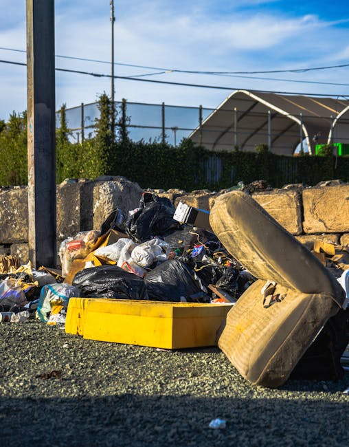 A collection of overflowing waste and recycling bins positioned on a paved sidewalk outside a commercial building with a construction scaffold in the background. The waste includes open cardboard boxes, plastic bags, and various packaging materials spilling onto the ground around the bins, which are mounted on a metal railing. The recycling containers are labeled for paper and mixed waste, while other bins are black and red for general waste. Several parked cars are visible behind the bins, and the environment is illuminated by daylight. This scene depicts the importance of proper waste disposal during home relocation and moving processes, as managed by professional removals services like Man with Van Primrose Hill.