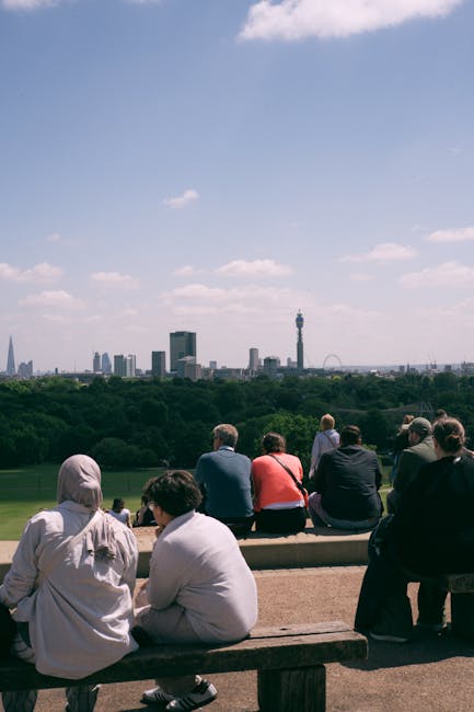 A group of people, including men and women, sitting on a wooden bench facing away from the camera, overlooks the London skyline from Primrose Hill on a clear day. Some individuals are dressed in casual clothing, with one woman wearing a headscarf. Behind them, a wide expanse of green parkland stretches out, with scattered trees and pathways. In the distance, tall city buildings, including a prominent TV tower and other skyscrapers, rise against a partly cloudy sky. The scene captures a peaceful moment of pedestrian enjoyment during a home relocation or moving activity in the area, with no visible furniture or packaging materials present.