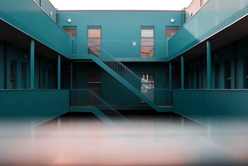 A multi-storey residential building featuring a turquoise exterior with metal railings and external staircases connecting the upper and lower levels. The building has several windows, some with curtains, and numbered apartment doors visible on the upper floor. The photo is taken from ground level, with a reflection slightly visible at the bottom of the image. This setting illustrates a typical environment where home relocation and furniture transport involve navigating staircases and parking areas, highlighting aspects essential for planning an efficient move. Man with Van Primrose Hill may use such properties for house removals, considering staircase access and parking options during the loading process of household items.