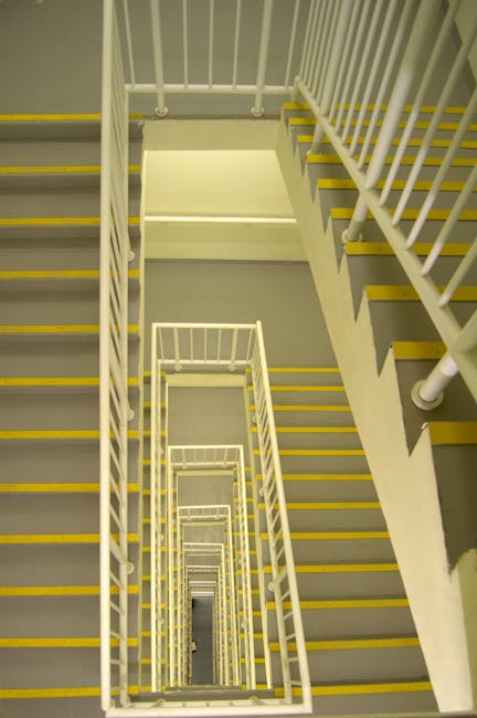 A view looking down a multi-story stairwell with white metal railings and yellow safety strips along the leading edges of each stair tread. The image captures the spiral descent from the top floor, showing the repeating pattern of stairs extending downward in a square configuration. The stairwell is illuminated by overhead lighting, casting consistent light across the white walls and railings. The photograph emphasizes the vertical movement involved in home relocation and furniture transport inside the building as part of a house removal process, with no objects or furniture visible but highlighting the typical stairwell environment encountered during domestic moves. The overall setting reflects the structural aspects of stair access used in building-wide moving logistics, maintained in a neutral and professional tone suitable for a moving services context.